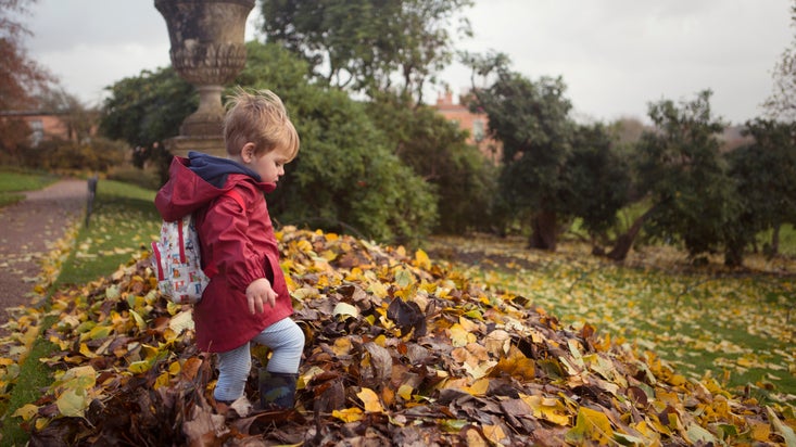 A child wearing a warm coat and boots kicks up a pile of leaves on the lawn, with an ornamental urn behind